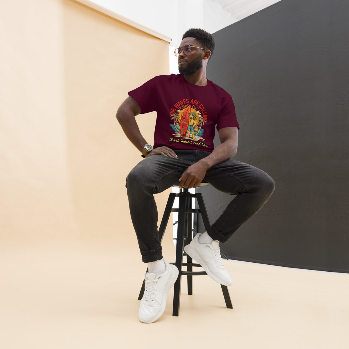Man wearing a maroon Lost Island Surf Club graphic T-shirt with a tropical island and surf scene, standing against a dark gray wall