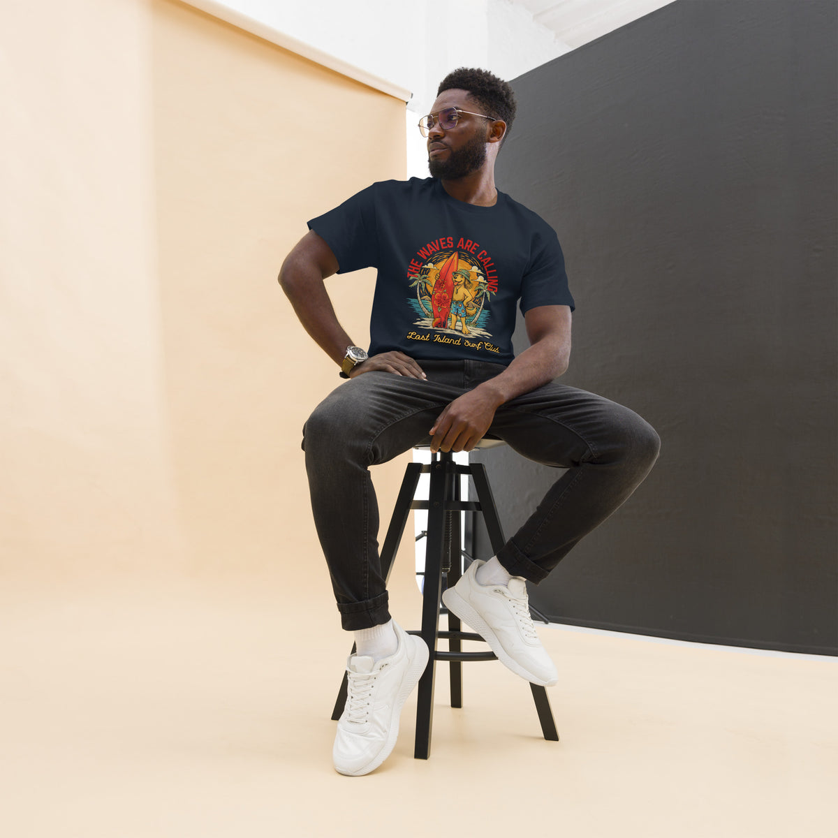 Man wearing a white Lost Island Surf Club graphic T-shirt with a colorful island scene, sitting on a stool with a beige background
